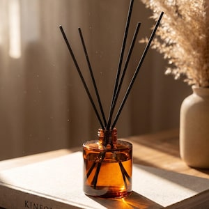 Peut inclure: Un diffuseur d'huiles essentielles en verre ambré avec des bâtonnets noirs, posé sur un livre avec le mot "KINFOLK" imprimé sur la couverture. Un vase avec des fleurs séchées est en arrière-plan. La scène est éclairée par la lumière naturelle.