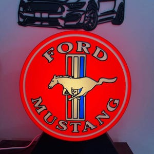 May include: A red Ford Mustang illuminated sign with a white running horse logo, and a black car silhouette above. The sign has a black base and a power cord.