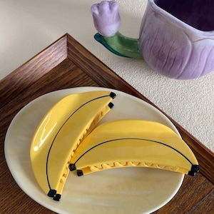 Könnte beinhalten: Zwei bananenförmige Haarspangen in Gelb mit schwarzen Details entlang der geschwungenen Kanten. Die Spangen haben eine glatte, glänzende Oberfläche und ein funktionales Krallendesign und liegen auf einem runden, cremefarbenen Teller.