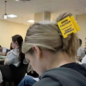 Könnte beinhalten: Eine Person mit blonden Haaren, die zu einem Dutt gebunden sind, trägt ein gelbes Schild mit der Aufschrift "CAUTION OVERSTIMULATED DONOT APPROACH". Die Person trägt einen grauen Kapuzenpullover und goldene Creolen. Andere Personen sind im Hintergrund zu sehen.