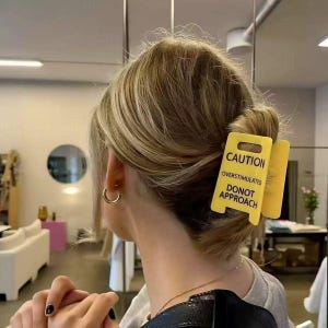 以下が含まれることがあります： 「CAUTION OVERSTIMULATED DONOT APPROACH」の黒い文字が入った、黄色の注意標識型のヘアクリップ。ブロンドヘアのお団子にクリップが留められています。ゴールドのフープイヤリングが見えます。