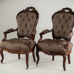May include: Two antique armchairs with dark brown wooden frames and tufted, brown fabric upholstery. The chairs have curved legs and armrests, and ornate carvings on the frames. The background is a plain white.