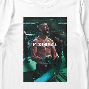 Pode incluir: Camiseta branca com um gráfico de um homem musculoso gritando, com o texto "F*CK THEM ALL" em branco. A imagem é uma fotografia de um lutador de UFC em uma arena de luta.