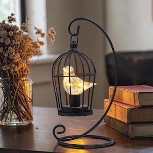 May include: A black metal birdcage-style lamp stand featuring a curved hook that suspends a small, clear glass bird figurine. The bird contains warm, glowing LED string lights inside, creating a soft illumination within the wire frame.