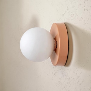 Peut inclure: Une applique murale composée d'un globe en verre blanc sphérique fixé sur une base circulaire de couleur pêche unie. Le luminaire s'étend vers l'extérieur depuis le mur, présentant un design lisse et arrondi avec une finition mate sur la base.