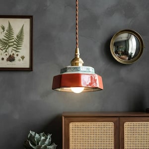 Puede incluir: Una lámpara colgante con una pantalla hexagonal de cerámica en color naranja quemado brillante, con un borde decorativo de patrones verdes y blancos. La pieza cuelga de un cable trenzado marrón e incluye un casquillo metálico dorado y una bombilla.