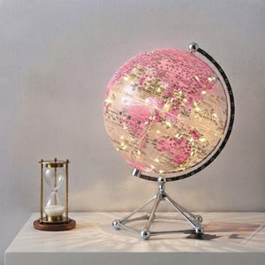 May include: A pink globe with black text and a silver meridian ring, illuminated by internal string lights, mounted on a silver tripod stand. Beside it sits a clear glass hourglass with a wooden base containing white sand.