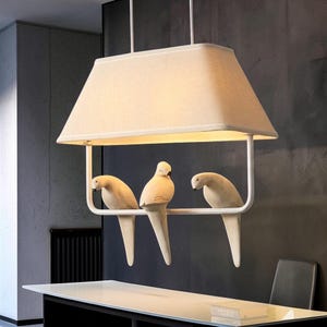 Może przedstawiać: Prostokątna lampa wisząca z beżowym abażurem z tkaniny zawieszonym na dwóch białych prętach. Pod abażurem znajduje się biała metalowa rama, na której w rzędzie siedzą trzy figurki ptaków w kolorze złamanej bieli, skierowane w dół i na boki.