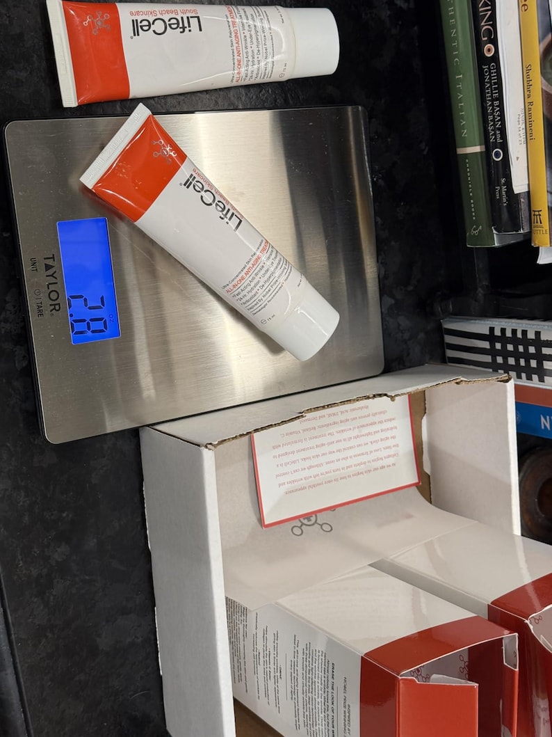 Pu&ograve; includere: Sono esposti due tubetti di crema anti-et&agrave; LifeCell con confezione rossa e bianca. Un tubetto poggia su una bilancia digitale, che mostra un peso di 2,8. La bilancia &egrave; argentata e lo sfondo include una scatola e libri parzialmente visibili.
