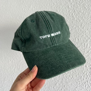 Könnte beinhalten: Eine dunkelgrüne Baseballkappe mit verwaschenem Vintage-Finish. Auf der Vorderseite ist der Schriftzug "TOFU MAUS" in weißem Garn eingestickt. Die Kappe verfügt über eine strukturierte Krone, einen gebogenen Schirm und gestickte Ösen.