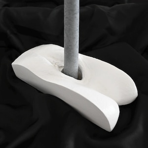 May include: A white, abstract sculpture with a curved, organic shape. A gray, cylindrical pole rises from a hole in the sculpture. The piece is set against a black background, highlighting its form and texture. The sculpture appears to be made of a solid, matte material.