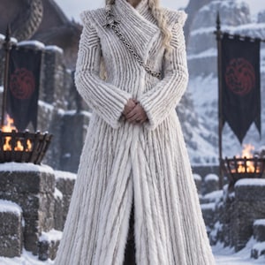 May include: A floor-length, off-white textured coat with a high collar and vertical ribbed detailing. The garment features a silver-toned metal chain draped diagonally across the chest, secured by a metallic dragon-shaped brooch at the shoulder.