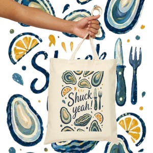 May include: A cream-colored tote bag with a design featuring oysters, lemon slices, and the phrase "Shuck yeah!". The bag is held by a person's hand against a white background with a repeating oyster pattern and cutlery.