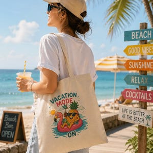 Può includere: Una borsa tote beige con una grafica colorata di un ananas con occhiali da sole che galleggia su un galleggiante a forma di fenicottero. Il testo "VACATION MODE ON" è sopra e sotto la grafica. La persona indossa una maglietta bianca, pantaloncini di jeans e un berretto beige.