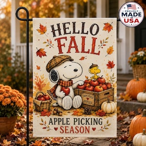 May include: A decorative garden flag featuring Snoopy and Woodstock with the text HELLO FALL and APPLE PICKING SEASON. The illustration shows Snoopy holding an apple next to a wooden crate labeled FARM FRESH APPLES, surrounded by autumn leaves.