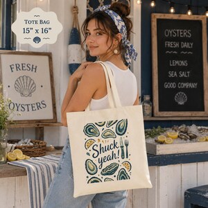 May include: Cream-colored tote bag, 15" x 16", with a design featuring oysters, lemon slices, and the phrase "Shuck yeah!". The bag has a seashell graphic and is held by a person. A sign in the background advertises fresh oysters.