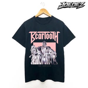 Puede incluir: Camiseta negra con el nombre de la banda "Beartooth" en letras rosas en bloque sobre un gráfico plateado y blanco. El texto "THE SURFACE" está debajo del gráfico. La marca "JOEDEZ" está en la esquina superior derecha.