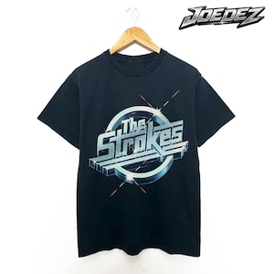 Puede incluir: Camiseta negra con el nombre de la banda "The Strokes" en un diseño 3D plateado y azul. La camiseta está colgada de una percha de madera sobre un fondo blanco. La marca "JOEDEZ" está impresa en la esquina superior derecha.