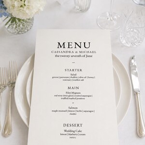 May include: A white rectangular wedding menu card with black serif text reading MENU, CASSANDRA & MICHAEL, the twenty seventh of June, STARTER, MAIN, and DESSERT, placed on a white dinner plate with silver forks and a knife on a light blue napkin.