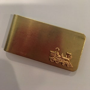 Könnte beinhalten: Ein rechteckiger Geldclip aus goldfarbenem Metall mit einer glatten, polierten Oberfläche. In der unteren rechten Ecke befindet sich ein kleines, detailliertes dreidimensionales Relief einer Dampflokomotive mit sichtbaren Rädern und Schornstein.