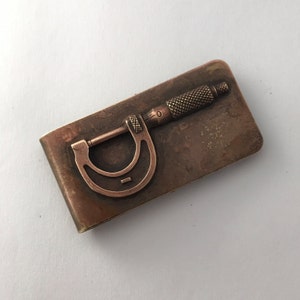 May include: A rectangular copper-toned money clip featuring a raised, three-dimensional relief of a micrometer tool. The metal surface has a weathered, oxidised patina with a textured finish, and the micrometer design includes a knurled grip.