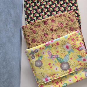 May include: Three fabric pieces with different patterns. The top piece has a black background with red cherries and white polka dots. The middle piece has a tan background with pink roses. The bottom piece has a yellow background with butterflies and flowers.