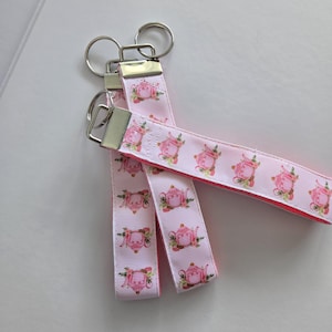 May include: Three keychains with pink ribbons featuring a repeating pattern of pink teapots with green leaves. The ribbons are attached to silver-colored key rings. The background is a light, neutral color.