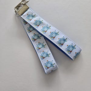 May include: A white ribbon keychain with a blue teapot pattern and a silver clasp. The ribbon has a blue border and is folded over. The teapot design includes blue, brown, and green accents.