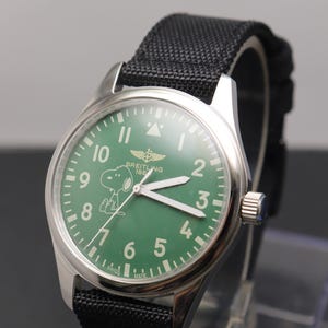 May include: A stainless steel wristwatch with a green dial featuring white Arabic numerals, a Snoopy character graphic, and the text Breitling 1884. The watch has white luminous hands, a silver crown, and a black textured fabric strap with stitching.