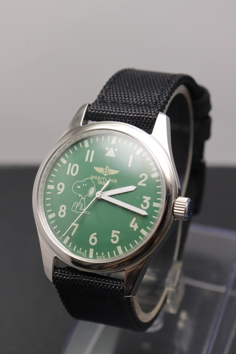May include: A stainless steel wristwatch with a green dial featuring white Arabic numerals, a Snoopy character graphic, and the text Breitling 1884. The watch has white luminous hands, a silver crown, and a black textured fabric strap with stitching.