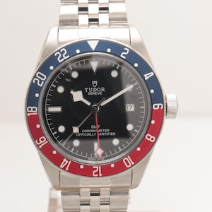 Puede incluir: Un reloj de pulsera Tudor GMT de acero inoxidable con esfera negra, marcadores de hora luminosos y una ventana de fecha en la posición de las tres. El reloj cuenta con un bisel giratorio bidireccional con inserto azul y rojo y brazalete metálico.