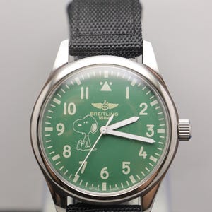 May include: A stainless steel wristwatch with a green dial featuring the character Snoopy, white Arabic numerals, and the text Breitling 1884. The watch has white luminous hands, a black fabric strap, and the text Swiss Made at the bottom.
