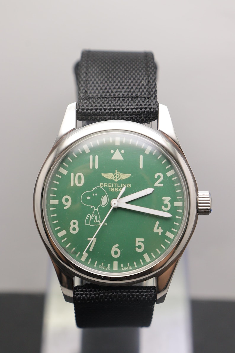 May include: A stainless steel wristwatch with a green dial featuring the character Snoopy, white Arabic numerals, and the text Breitling 1884. The watch has white luminous hands, a black fabric strap, and the text Swiss Made at the bottom.