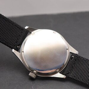 May include: A silver-toned wristwatch with a plain, circular metal case back and a textured black nylon strap. The watch features a small, ridged winding crown positioned on the side of the case, attached to the lugs holding the black fabric band.