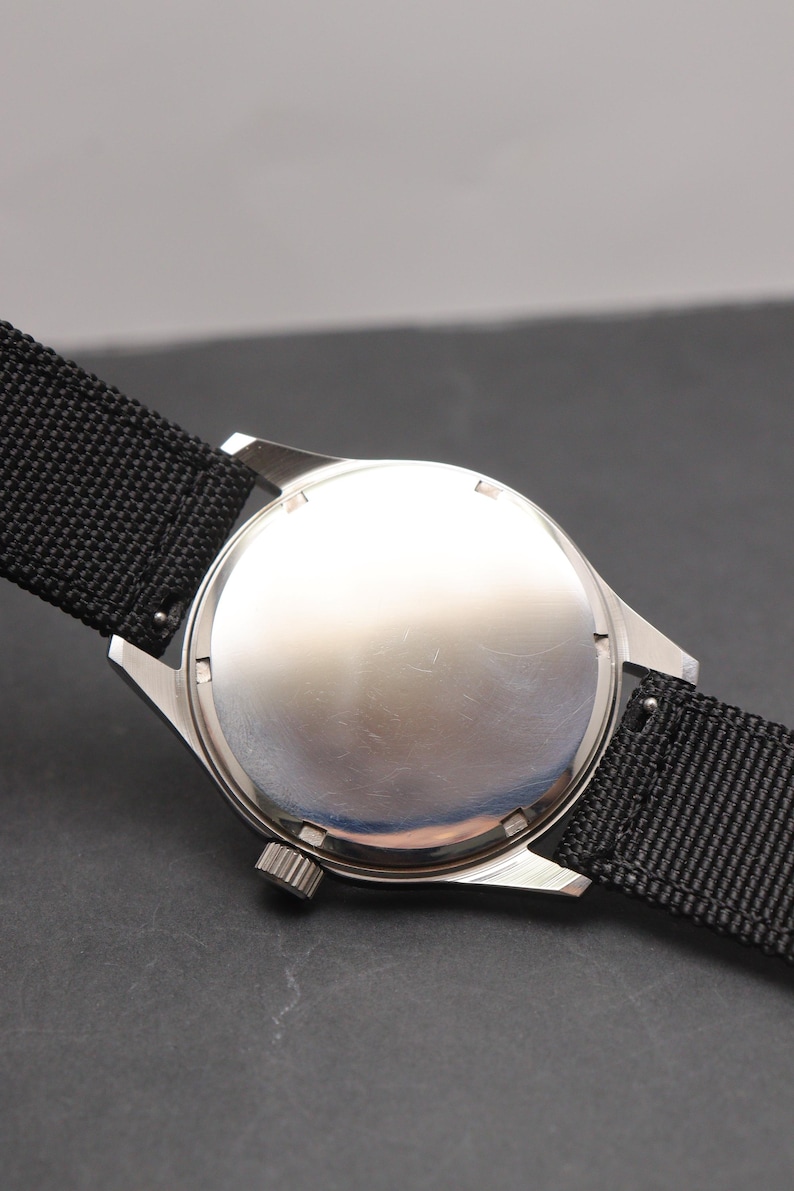 May include: A silver-toned wristwatch with a plain, circular metal case back and a textured black nylon strap. The watch features a small, ridged winding crown positioned on the side of the case, attached to the lugs holding the black fabric band.