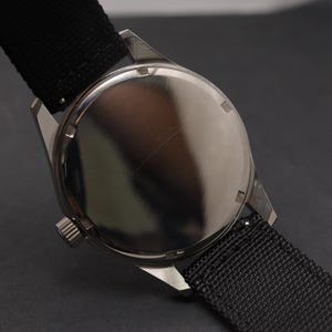 May include: A wristwatch with a polished silver-tone circular case back and a black textured nylon strap. The watch features a small, ridged crown on the side of the case, and the metal back has a smooth, reflective surface with visible light reflections.