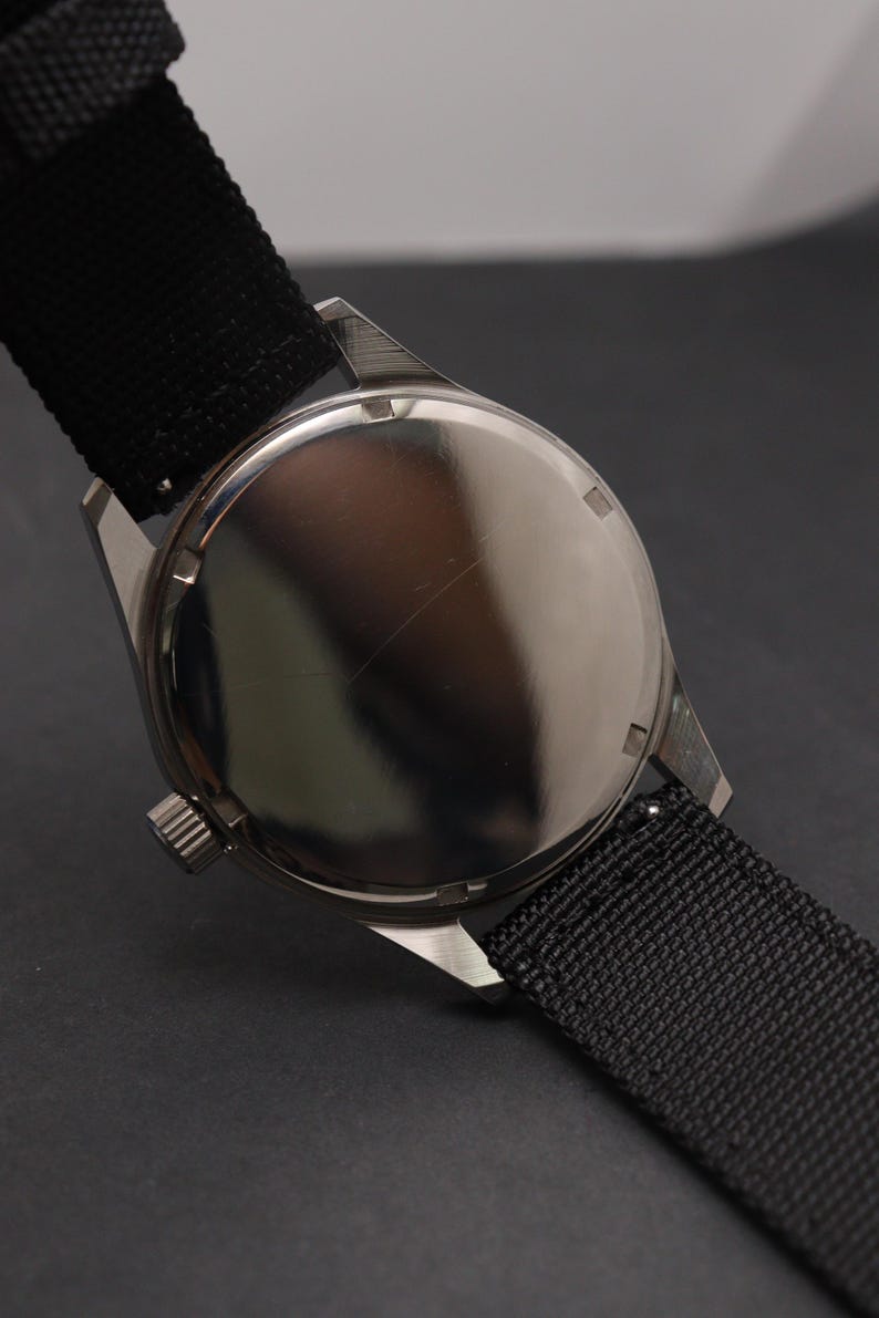 May include: A wristwatch with a polished silver-tone circular case back and a black textured nylon strap. The watch features a small, ridged crown on the side of the case, and the metal back has a smooth, reflective surface with visible light reflections.