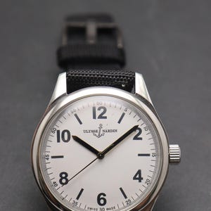 May include: A silver-toned wristwatch with a white dial, black Arabic numerals at the 2, 4, 6, 8, 10, and 12 positions, and black hands. The dial features the text Ulysse Nardin with an anchor logo and Swiss Made at the bottom. A black strap is attached.