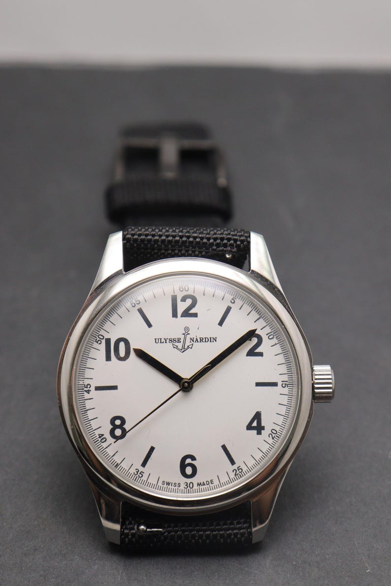 May include: A silver-toned wristwatch with a white dial, black Arabic numerals at the 2, 4, 6, 8, 10, and 12 positions, and black hands. The dial features the text Ulysse Nardin with an anchor logo and Swiss Made at the bottom. A black strap is attached.