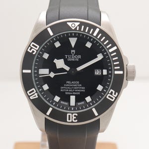 Op de afbeelding: Een Tudor Pelagos polshorloge met een zwarte wijzerplaat, witte lichtgevende uurmarkeringen en een datumvenster op de drie-uur-positie. Het horloge heeft een zwarte draaibare ring met witte cijfers en een grijze rubberen horlogeband.