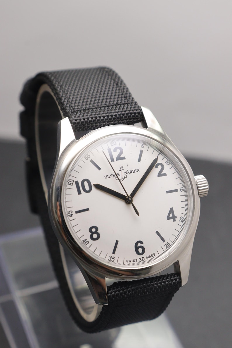 May include: A silver-toned wristwatch with a white dial, black Arabic numerals, and black hands. The dial features the text ULYSSE NARDIN with an anchor logo and SWISS MADE at the bottom. It has a black textured fabric strap with a silver buckle.
