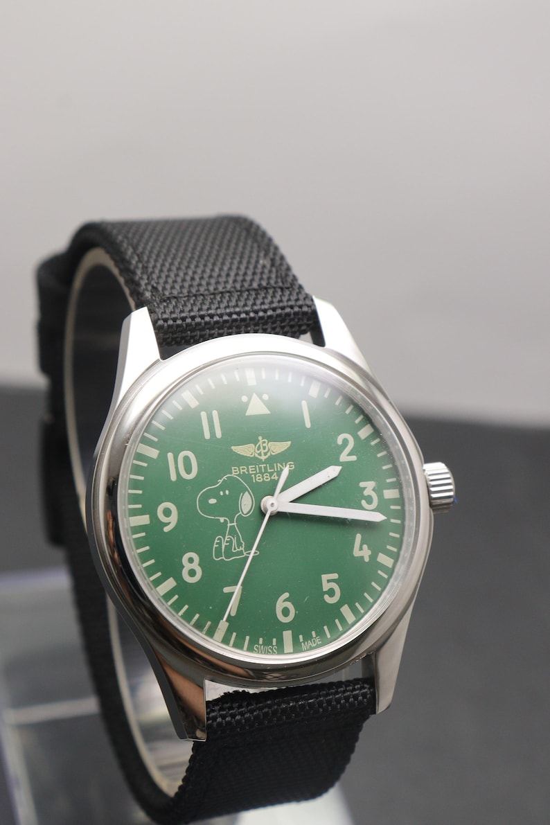 May include: A silver-tone wristwatch with a green dial featuring white Arabic numerals, a Snoopy character graphic, and the text Breitling 1884. The watch has a black textured fabric strap and white hour, minute, and second hands.