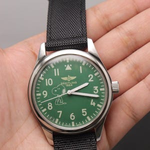 May include: A stainless steel wristwatch with a green dial featuring white Arabic numerals, a Breitling 1884 logo, and a graphic of Snoopy. The watch has white hour and minute hands, a black fabric strap, and the text Swiss Made at the bottom.