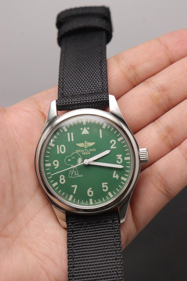 May include: A stainless steel wristwatch with a green dial featuring white Arabic numerals, a Breitling 1884 logo, and a graphic of Snoopy. The watch has white hour and minute hands, a black fabric strap, and the text Swiss Made at the bottom.