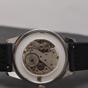 May include: A silver-toned wristwatch with an exposed mechanical movement featuring seventeen jewels and Swiss markings. The watch has a white inner ring, a metallic gear mechanism, and a black textured fabric strap attached to the lugs.