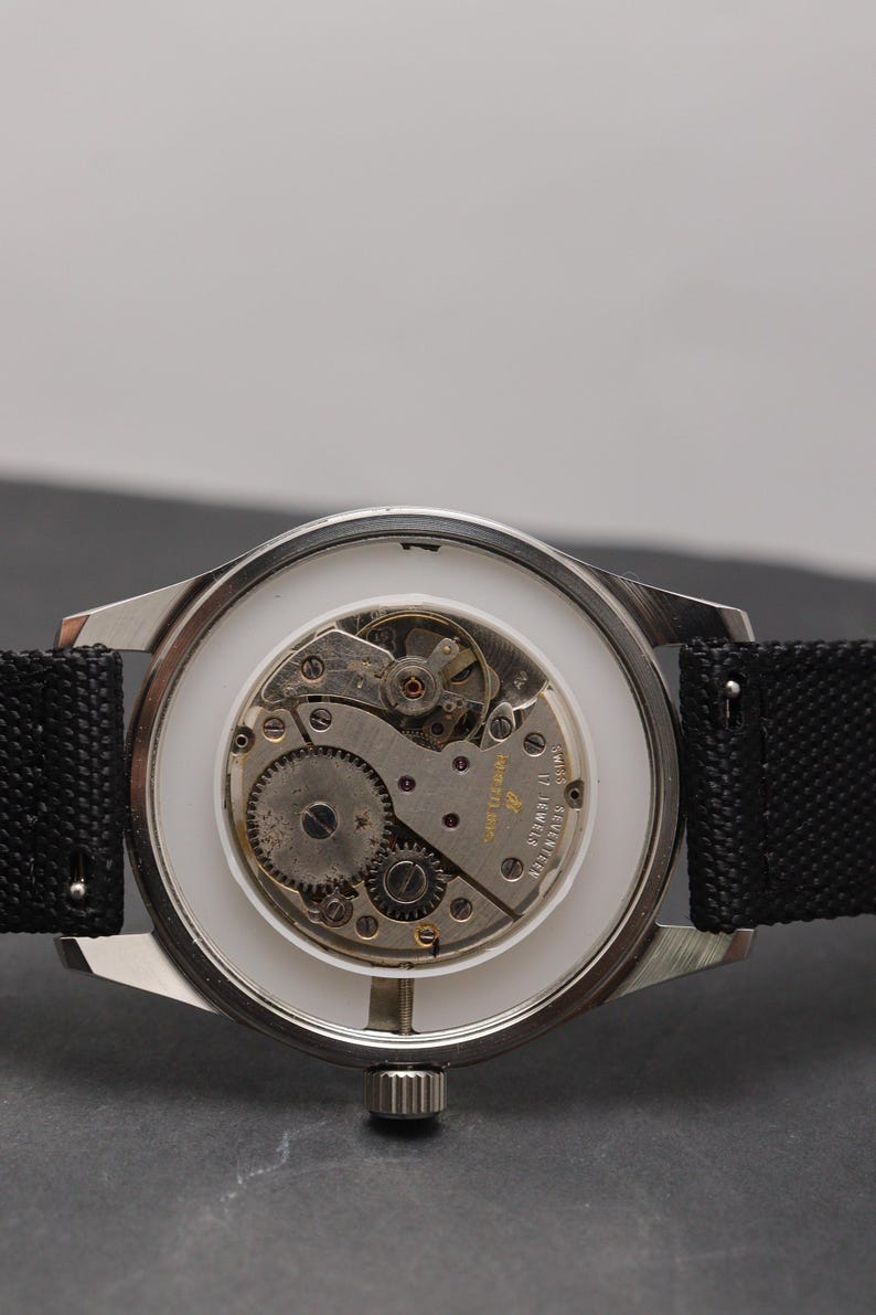 May include: A silver-toned wristwatch with an exposed mechanical movement featuring seventeen jewels and Swiss markings. The watch has a white inner ring, a metallic gear mechanism, and a black textured fabric strap attached to the lugs.
