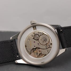 May include: A silver-toned wristwatch with an exposed mechanical movement visible through the back casing. The watch features a black textured fabric strap, a white inner ring, and a metallic crown positioned at the top of the circular case.