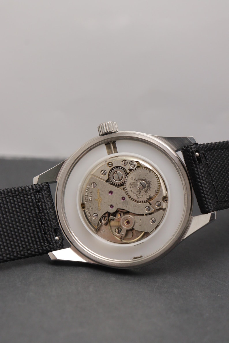 May include: A silver-toned wristwatch with an exposed mechanical movement visible through the back casing. The watch features a black textured fabric strap, a white inner ring, and a metallic crown positioned at the top of the circular case.