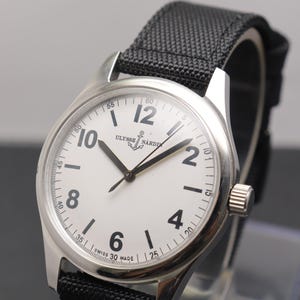 May include: A stainless steel wristwatch with a white dial, black Arabic numerals, and a black textured fabric strap. The dial features the Ulysse Nardin logo with an anchor symbol and the text Swiss Made at the bottom. The watch has black hands.