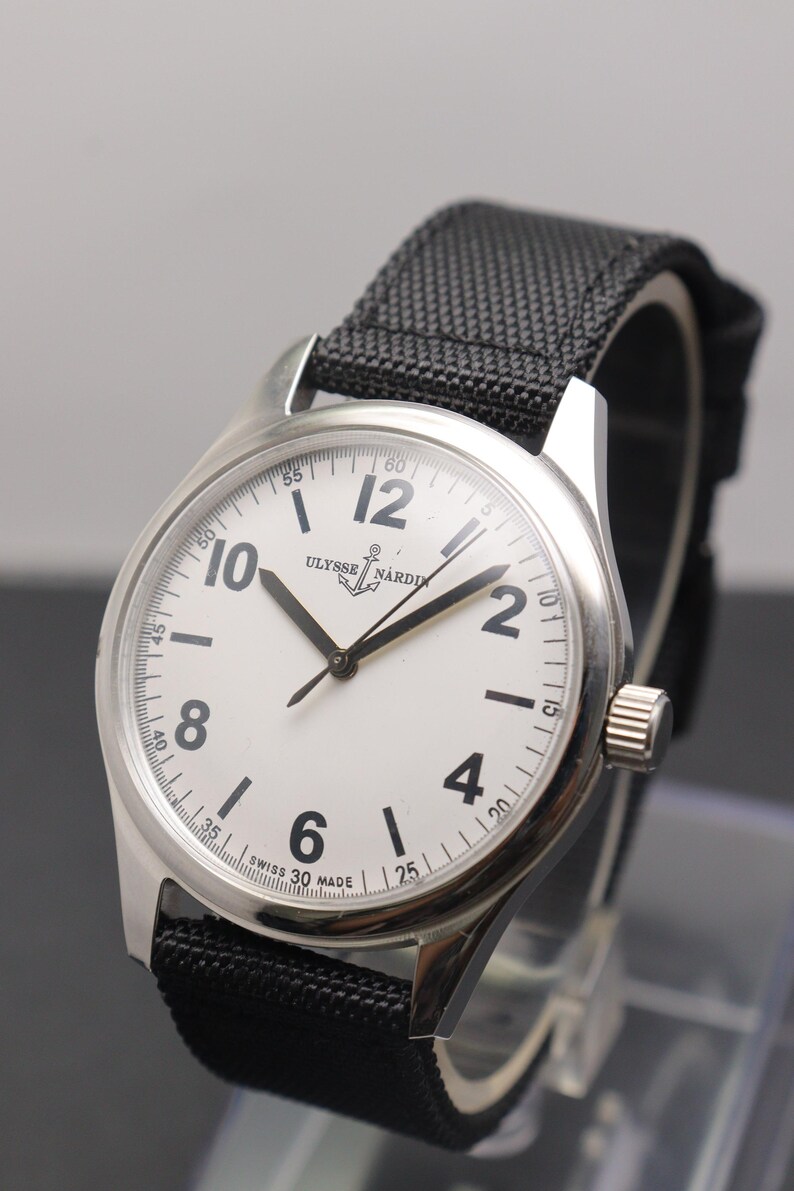 May include: A stainless steel wristwatch with a white dial, black Arabic numerals, and a black textured fabric strap. The dial features the Ulysse Nardin logo with an anchor symbol and the text Swiss Made at the bottom. The watch has black hands.